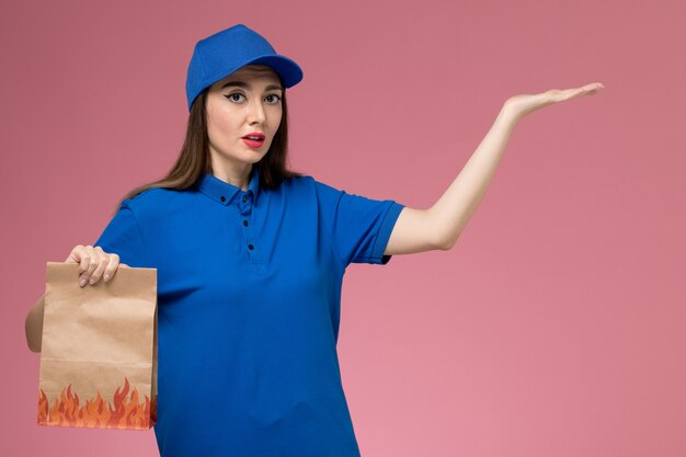 Front view female courier in blue uniform and cape holding paper food package smiling on the pink desk 