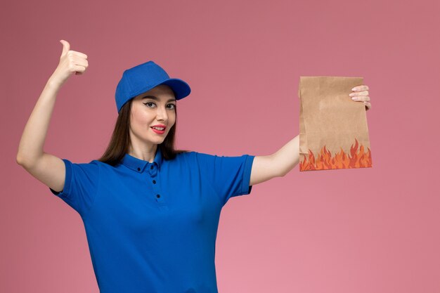 파란색 유니폼과 케이프 핑크 벽에 미소 종이 음식 패키지를 들고 전면보기 여성 택배