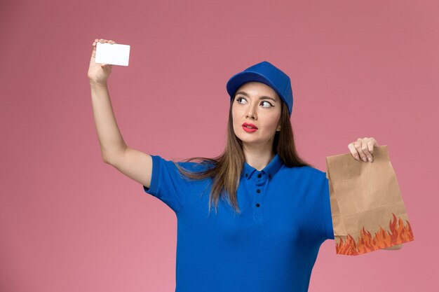 Front view female courier in blue uniform and cape holding white card and paper food package on pink wall 