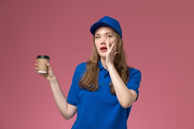 Вид спереди женщина-курьер в синей форме, держащая коричневую кофейную чашку на светло-розовом столе, сервисная рабочая форма, доставка компании