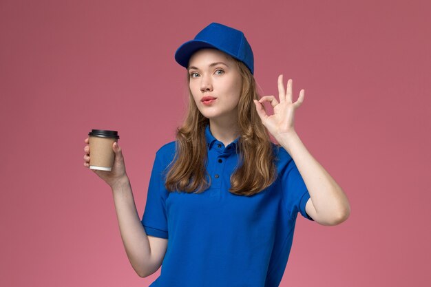 Front view female courier in blue uniform holding brown coffee cup on the light-pink desk service uniform delivering company job work