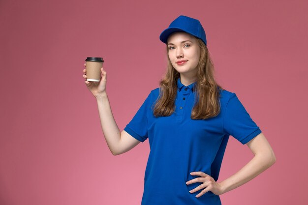 Front view female courier in blue uniform holding brown coffee cup on pink desk service uniform delivering company job work