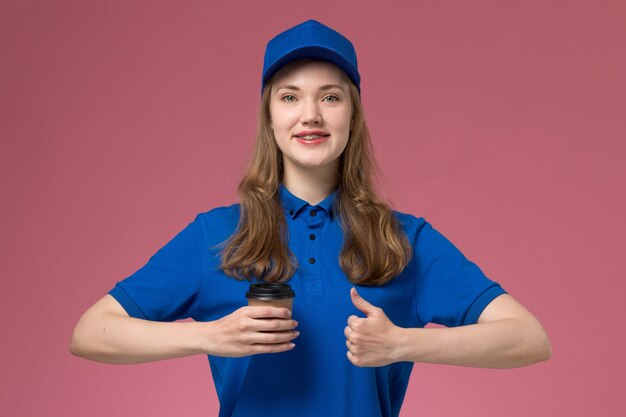 Front view female courier in blue uniform holding brown coffee cup on the pink desk service uniform delivering company job work