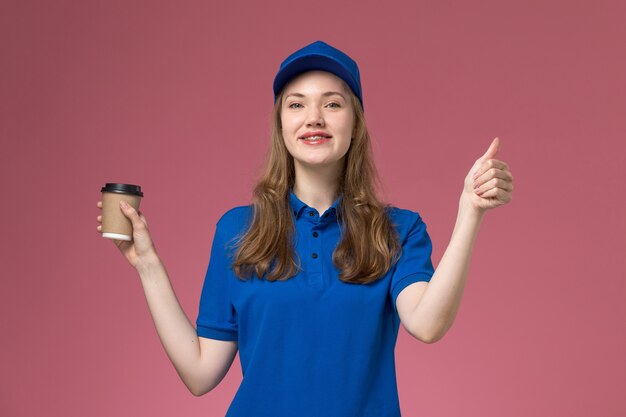 Front view female courier in blue uniform holding brown coffee cup smiling on the light-pink desk service uniform delivering company job work