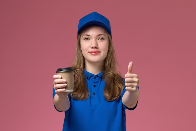 Front view female courier in blue uniform holding brown coffee cup smiling on pink desk service uniform delivering company job work