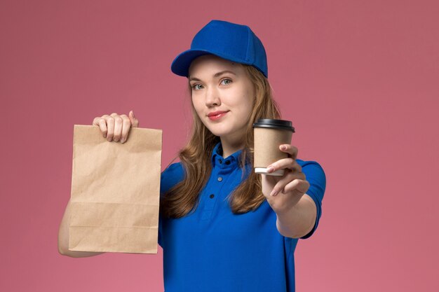Вид спереди курьер-женщина в синей форме, держащая коричневую кофейную чашку с пакетом продуктов и улыбку на розовом столе, обслуживающая униформу, доставка компании