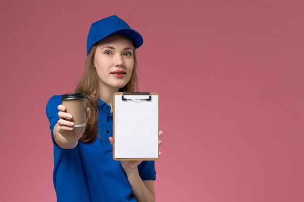 Женщина-курьер в синей униформе, держащая коричневую кофейную чашку с блокнотом на светло-розовом столе, служба доставки униформы, вид спереди