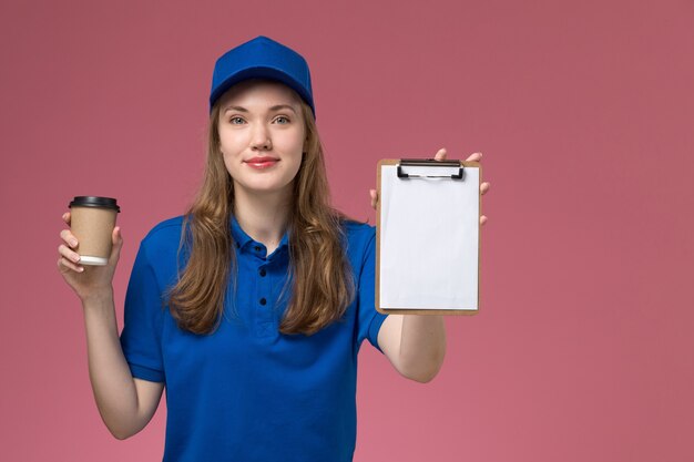 Front view female courier in blue uniform holding brown coffee cup with notepad on pink background service job uniform work delivering company