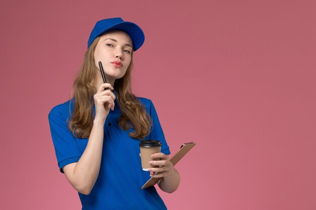 正面図青い制服を着た女性の宅配便は、メモ帳付きの茶色のコーヒーカップを保持し、淡いピンクのデスクサービス制服配達会社で表現を考えています