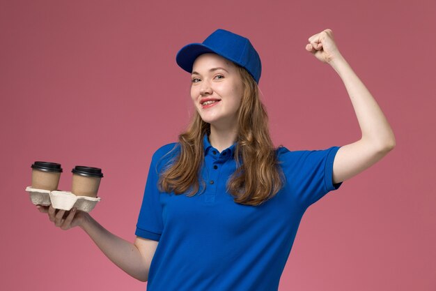 Front view female courier in blue uniform holding brown coffee cups flexing with smile on the pink background service uniform delivering company job