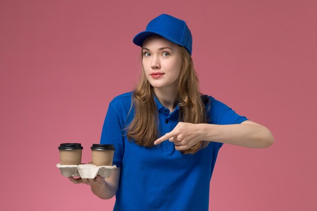 Женщина-курьер в синей форме, держащая коричневые кофейные чашки на розовом столе, служба доставки