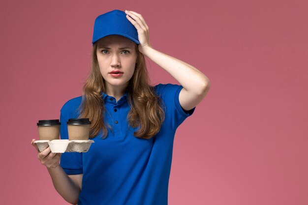 Front view female courier in blue uniform holding brown coffee cups on pink floor service uniform delivering company job