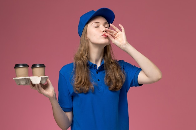 ピンクの背景に味のサインを示す茶色のコーヒーカップを保持している青い制服の正面図女性宅配便サービス制服配達会社の仕事