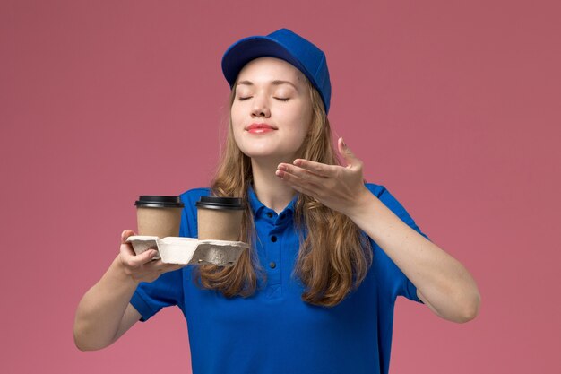 ピンクの背景のサービスユニフォームでそれらをかぐ茶色のコーヒーカップを保持している青い制服の正面図の女性の宅配便会社の仕事を提供します