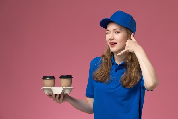 회사 작업을 제공하는 핑크 데스크 서비스 유니폼에 윙크하는 showign 전화 제스처를 윙크하는 갈색 커피 컵을 들고 파란색 유니폼에 전면보기 여성 택배