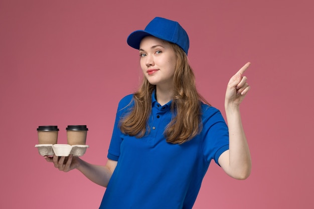 Вид спереди женщина-курьер в синей форме, держащая коричневые кофейные чашки с легкой улыбкой на розовом фоне, сервисная форма, доставляющая работу компании