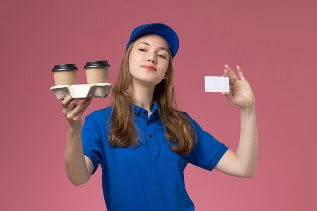 Front view female courier in blue uniform holding brown delivery coffee cups and card on the pink background service uniform delivering company job