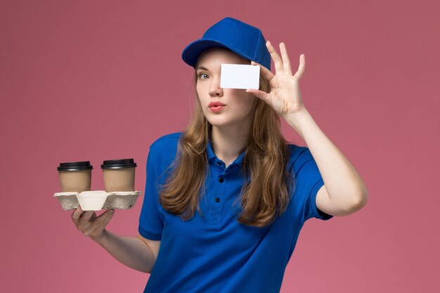 Front view female courier in blue uniform holding brown delivery coffee cups and card on the pink desk service uniform delivering company job