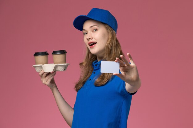 Front view female courier in blue uniform holding brown delivery coffee cups and card on pink desk service uniform delivering company