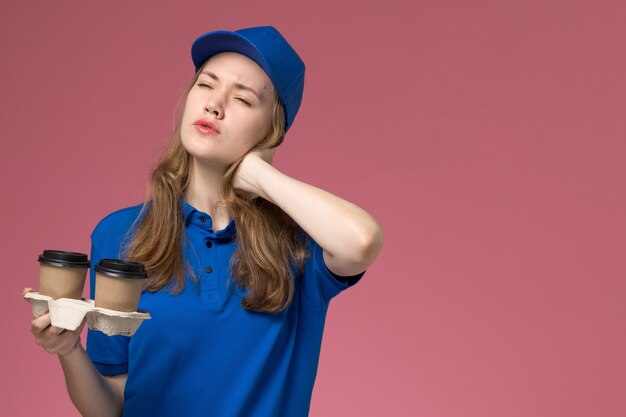 Front view female courier in blue uniform holding brown delivery coffee cups having neck ache on pink background service uniform delivering company job