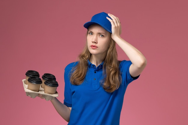 Front view female courier in blue uniform holding brown delivery coffee cups holding her head on pink desk service uniform company worker