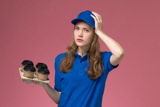Front view female courier in blue uniform holding brown delivery coffee cups holding her head on pink desk service uniform company worker