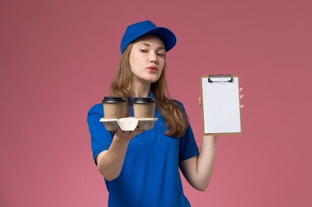 Front view female courier in blue uniform holding brown delivery coffee cups and notepad on pink desk service uniform company