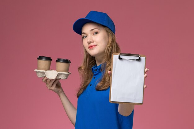 ピンクのデスクサービスの仕事の制服会社の仕事に笑顔で茶色の配達コーヒーカップとメモ帳を保持している青い制服の正面図女性宅配便