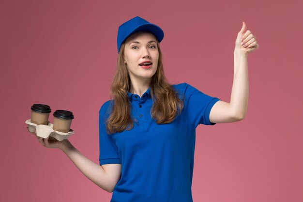 Вид спереди женщина-курьер в синей форме, держащая коричневые кофейные чашки доставки на розовом фоне, служебная форма, выполняющая работу компании