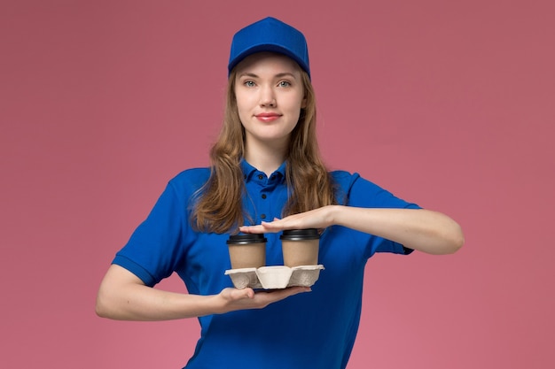Вид спереди женщина-курьер в синей форме, держащая коричневые чашки кофе с доставкой и улыбающаяся на розовом фоне, сервисная форма, доставляющая работу компании