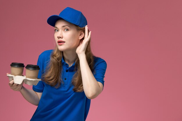 회사 작업을 제공하는 분홍색 배경 서비스 제복에 귀를 기울이려고 갈색 배달 커피 컵을 들고 파란색 제복을 입은 전면보기 여성 택배