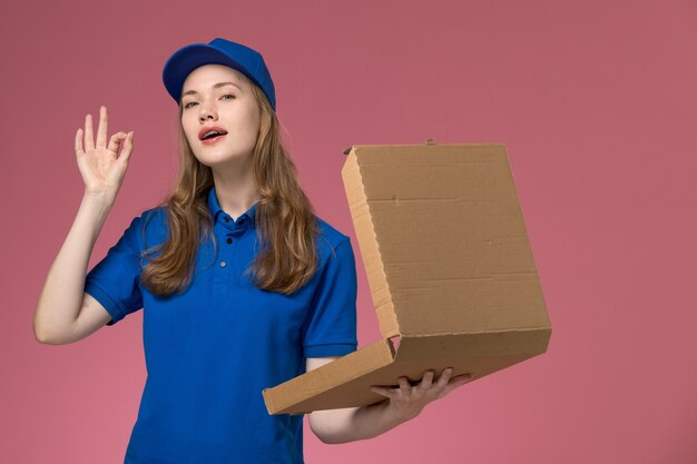 Вид спереди женщина-курьер в синей униформе держит пустую коробку для доставки еды на розовом столе.