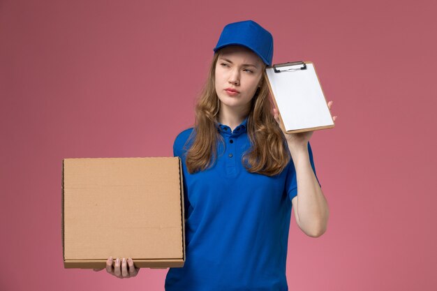 Front view female courier in blue uniform holding food delivery box and notepad on the light-pink desk service uniform job company worker