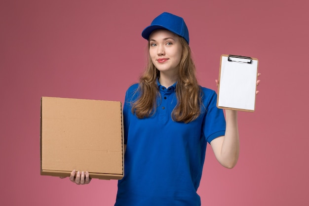 Вид спереди женщина-курьер в синей форме, держащая коробку для доставки еды и блокнот на розовом столе, служебная форма, работник компании