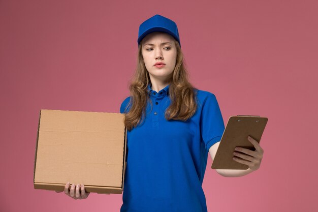 Foto gratuita corriere femminile di vista frontale in uniforme blu che tiene scatola di consegna di cibo e blocco note con espressione confusa sulla società di lavoro uniforme servizio scrivania rosa