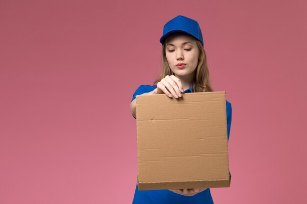 Front view female courier in blue uniform holding food delivery box opening it on pink background job service uniform company