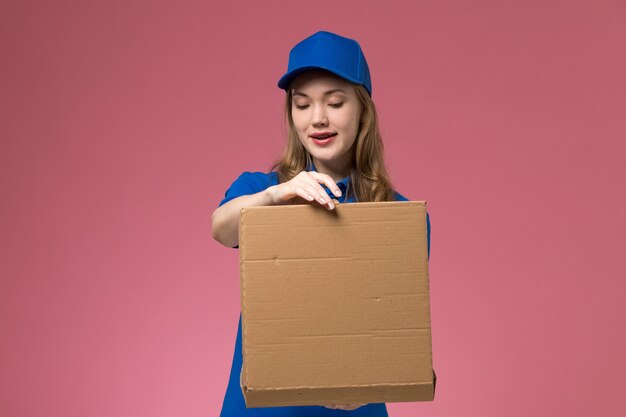 ピンクのデスクの仕事サービスの制服会社でそれを開く食品配達ボックスを保持している青い制服の正面図女性宅配便