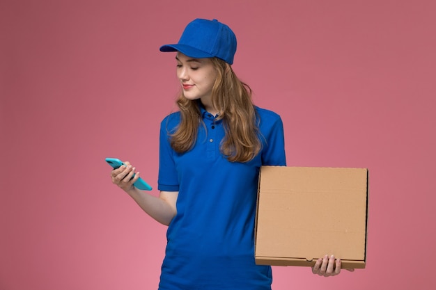 Вид спереди женщина-курьер в синей форме с коробкой для доставки еды и телефоном на розовом столе.