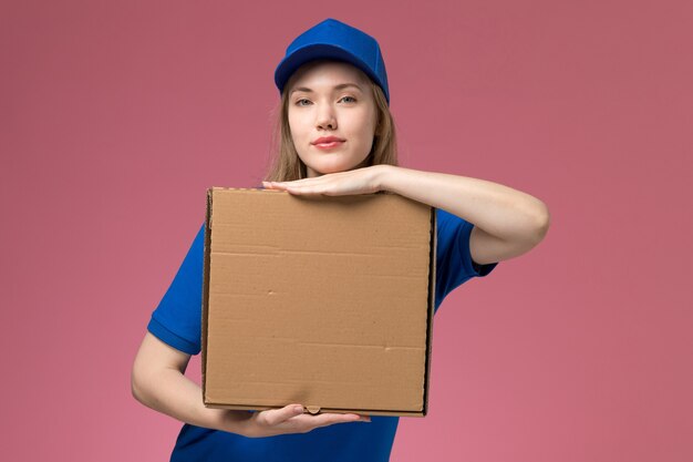 Вид спереди женщина-курьер в синей форме с коробкой для доставки еды на розовом фоне.