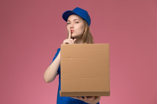 Вид спереди женщина-курьер в синей форме, держащая коробку для доставки еды, показывая знак тишины на розовом столе, служба работы, униформа, компания