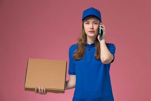 Front view female courier in blue uniform holding food delivery box and talking on the phone on pink background service uniform company job