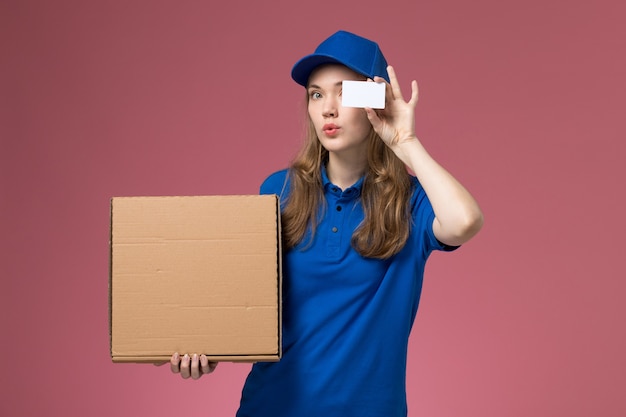 Вид спереди женщина-курьер в синей форме, держащая коробку для доставки еды и белую карточку на розовом столе, сервисная форма, работник компании