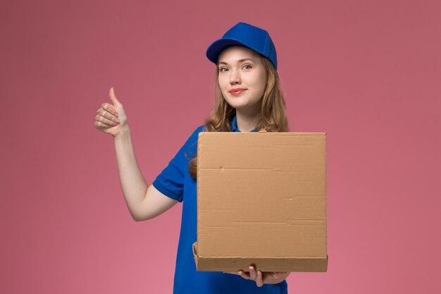 Вид спереди женский курьер в синей форме, держащий коробку для доставки еды с улыбкой на розовом фоне, служба униформы компании