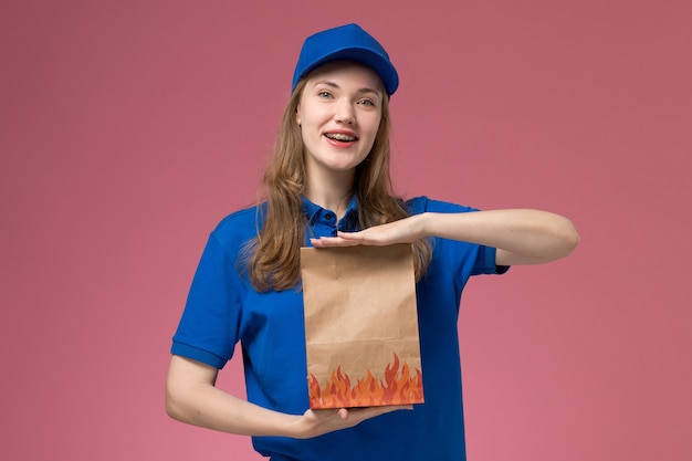 Front view female courier in blue uniform holding food package smiling on pink background job worker service uniform company