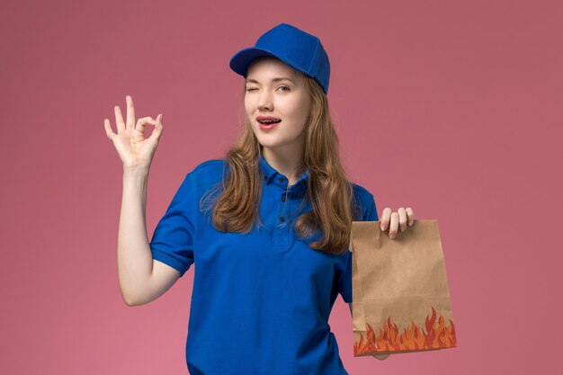 Вид спереди женщина-курьер в синей форме, держащая продуктовый пакет, улыбается и подмигивает светло-розовым рабочим столом службы униформы компании