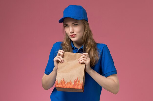 Front view female courier in blue uniform holding food package with skeptical expression on the pink background job worker service uniform company
