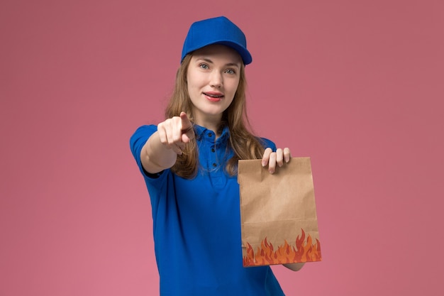 Foto gratuita corriere femminile di vista frontale in uniforme blu che tiene il pacchetto alimentare con un leggero sorriso sottolineando sulla società uniforme di servizio di lavoro scrivania rosa chiaro