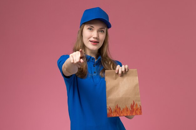 Front view female courier in blue uniform holding food package with slight smile pointing out on light pink desk job worker service uniform company