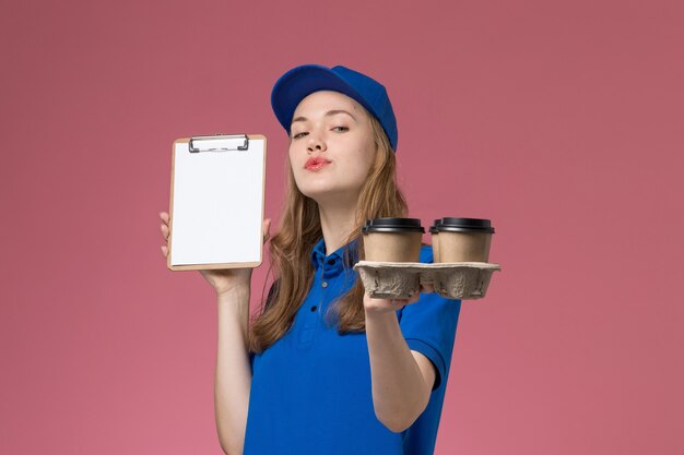 Foto gratuita corriere femminile di vista frontale in uniforme blu che tiene il blocco note e le tazze di caffè di consegna sul lavoro dell'azienda dell'uniforme di servizio della reception rosa-chiaro