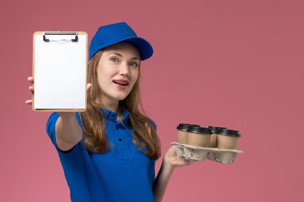 Front view female courier in blue uniform holding notepad and delivery coffee cups on pink light desk service uniform company